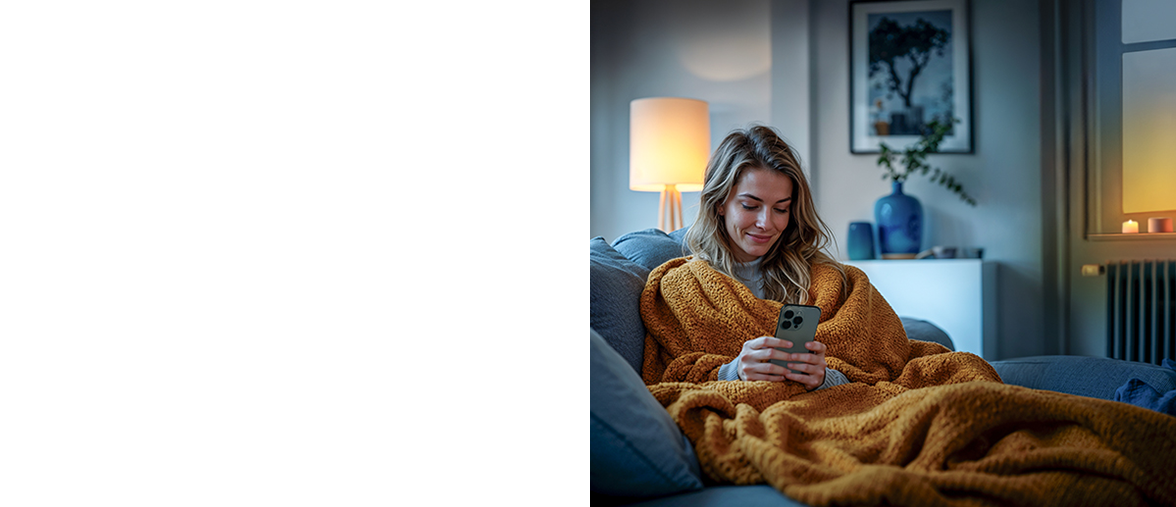A woman sits snuggled up in a blanket on the sofa and operates her lamps and heating thermostats using the Hama app.